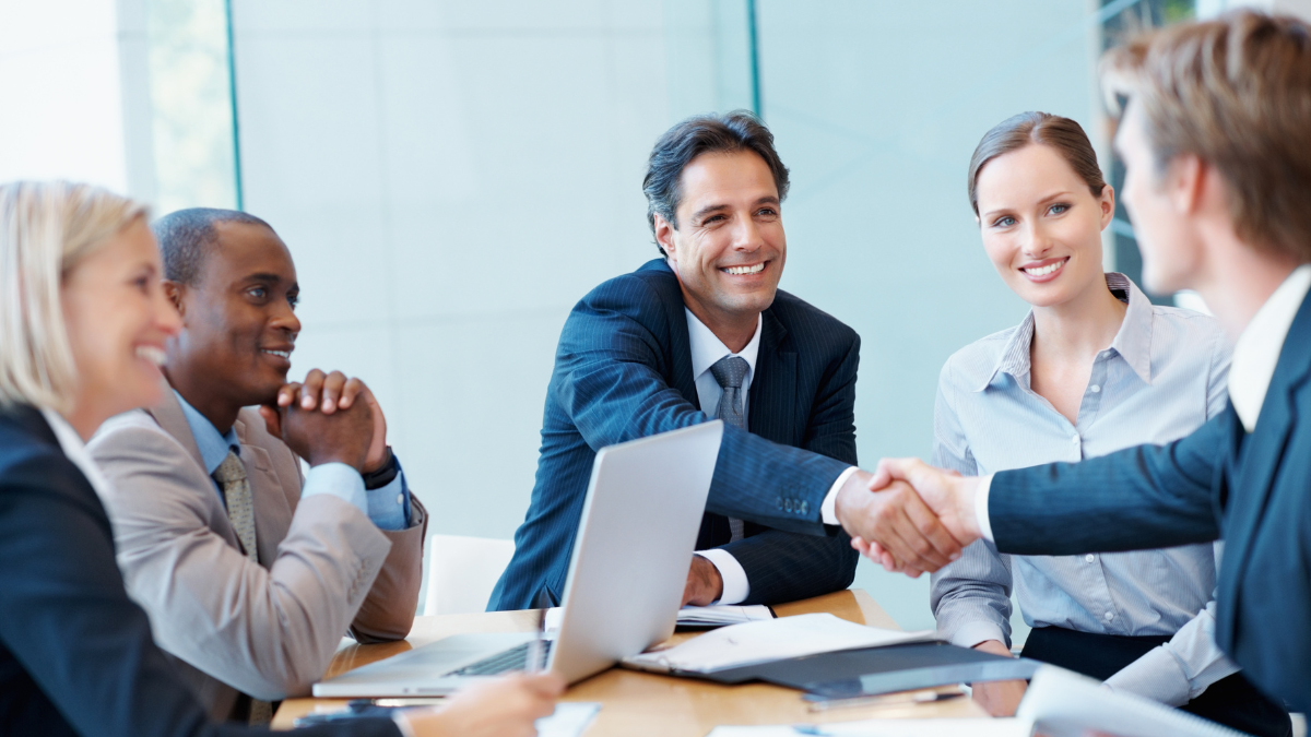 Group of professionals in a meeting, with two individuals shaking hands across the table.