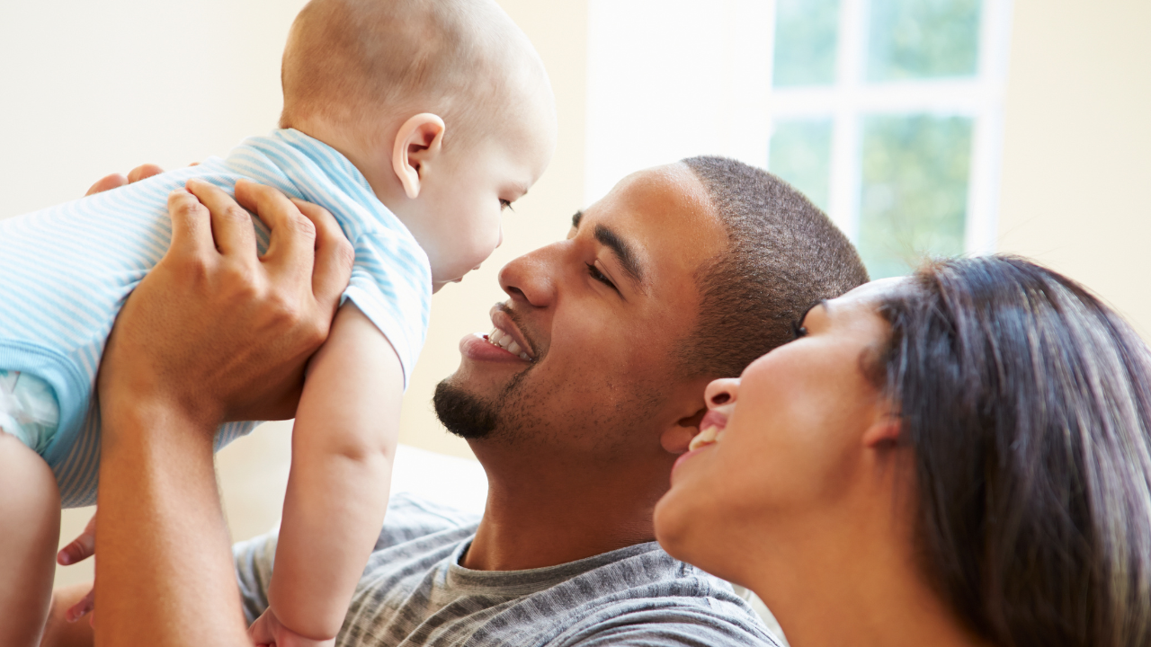 Smiling parents interacting with a baby in a bright, homey space.